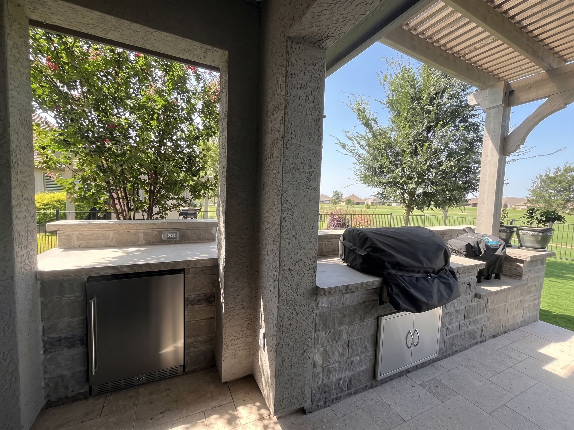 Covered outdoor patio with grill, sink, and stone countertop beside a yard and trees