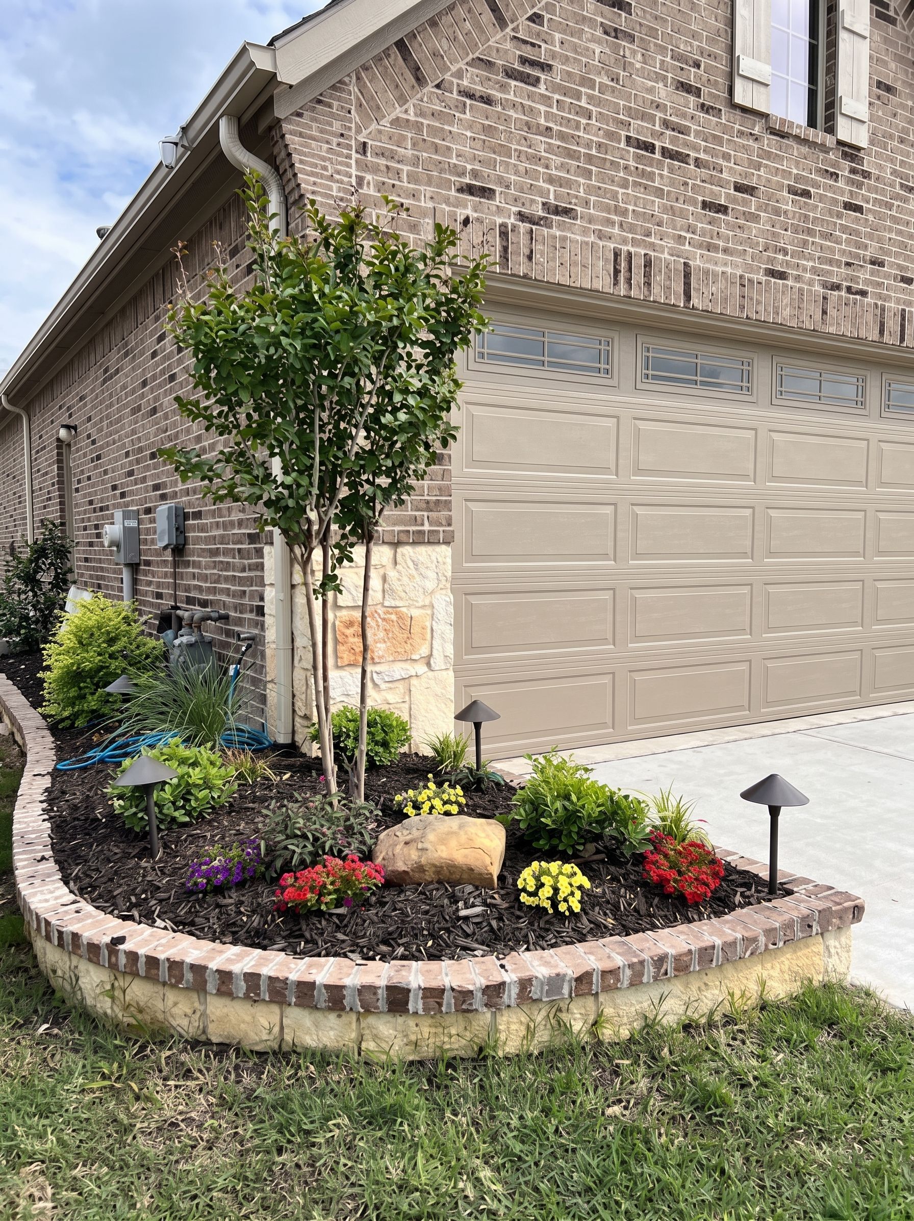 Brick house corner with garage, landscaped flower bed, small tree, and lawn