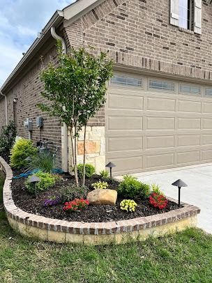 Brick house corner with flower bed, young tree, and garage door