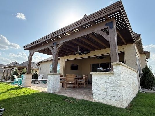 Covered patio on a beige house with wood beams, stone columns, outdoor dining set, and green lawn