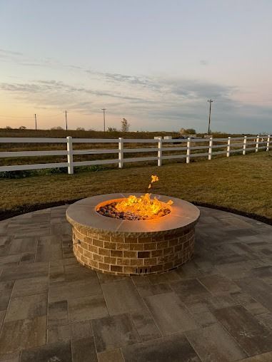 Round brick fire pit burning on patio beside white fence at sunset
