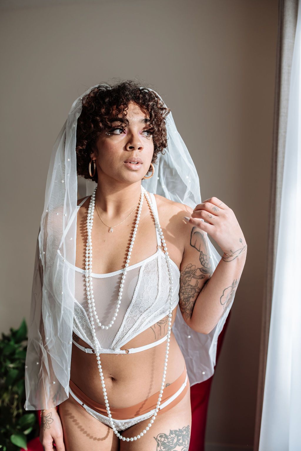 A woman in lingerie and a veil is standing in front of a window.
