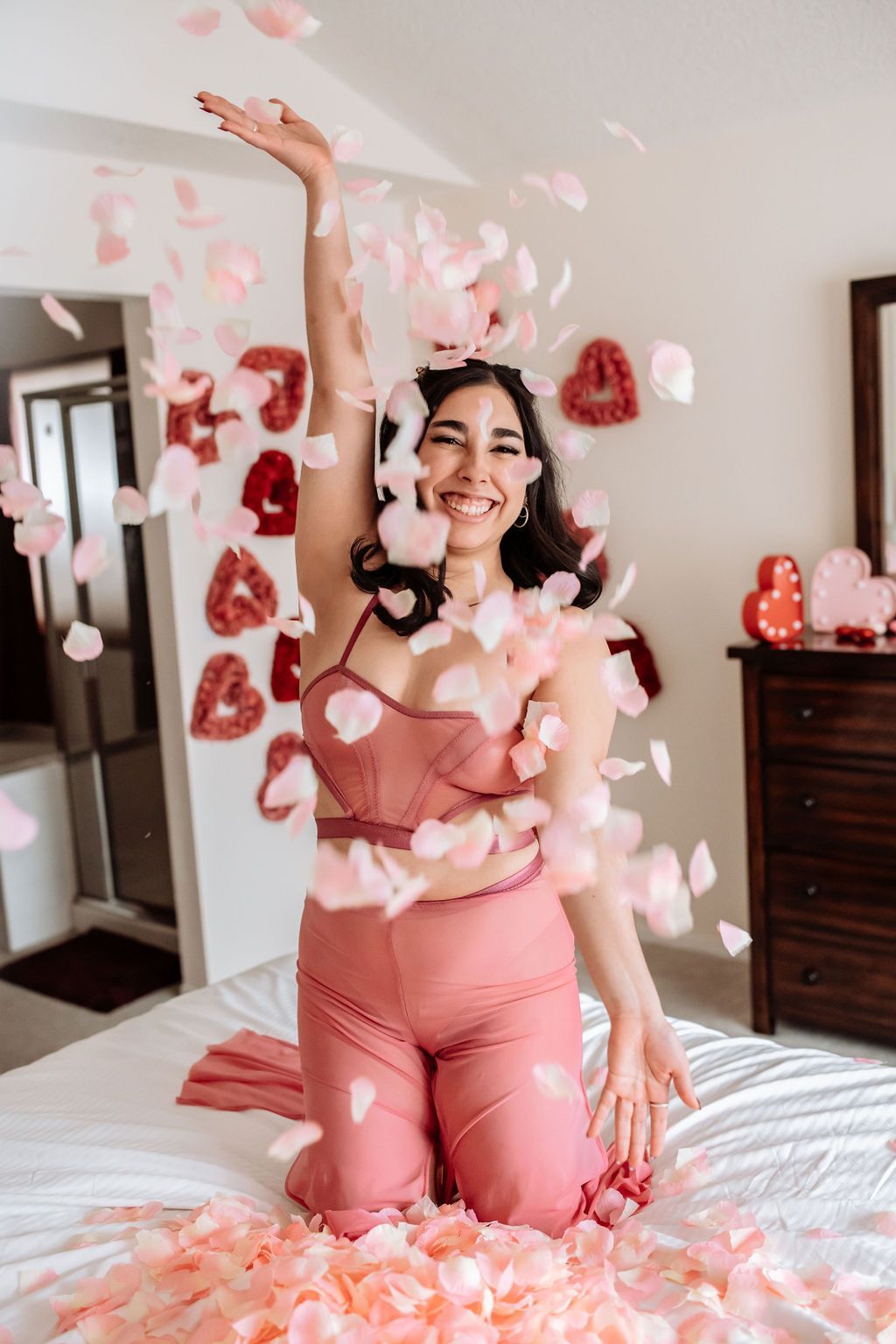 A woman is kneeling on a bed with petals falling from the ceiling.
