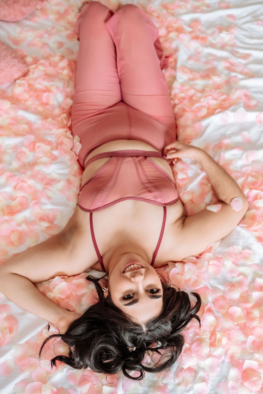 looking down on a woman laying down on a bed that is covered in petals.  She is wearing a pink bralette and pink pants.