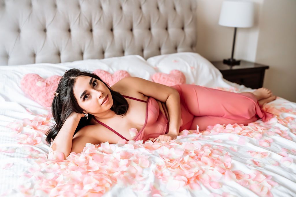 A woman laying on her side on a bed covered in petals.  She is wearing a pink bralette and matching pants. 