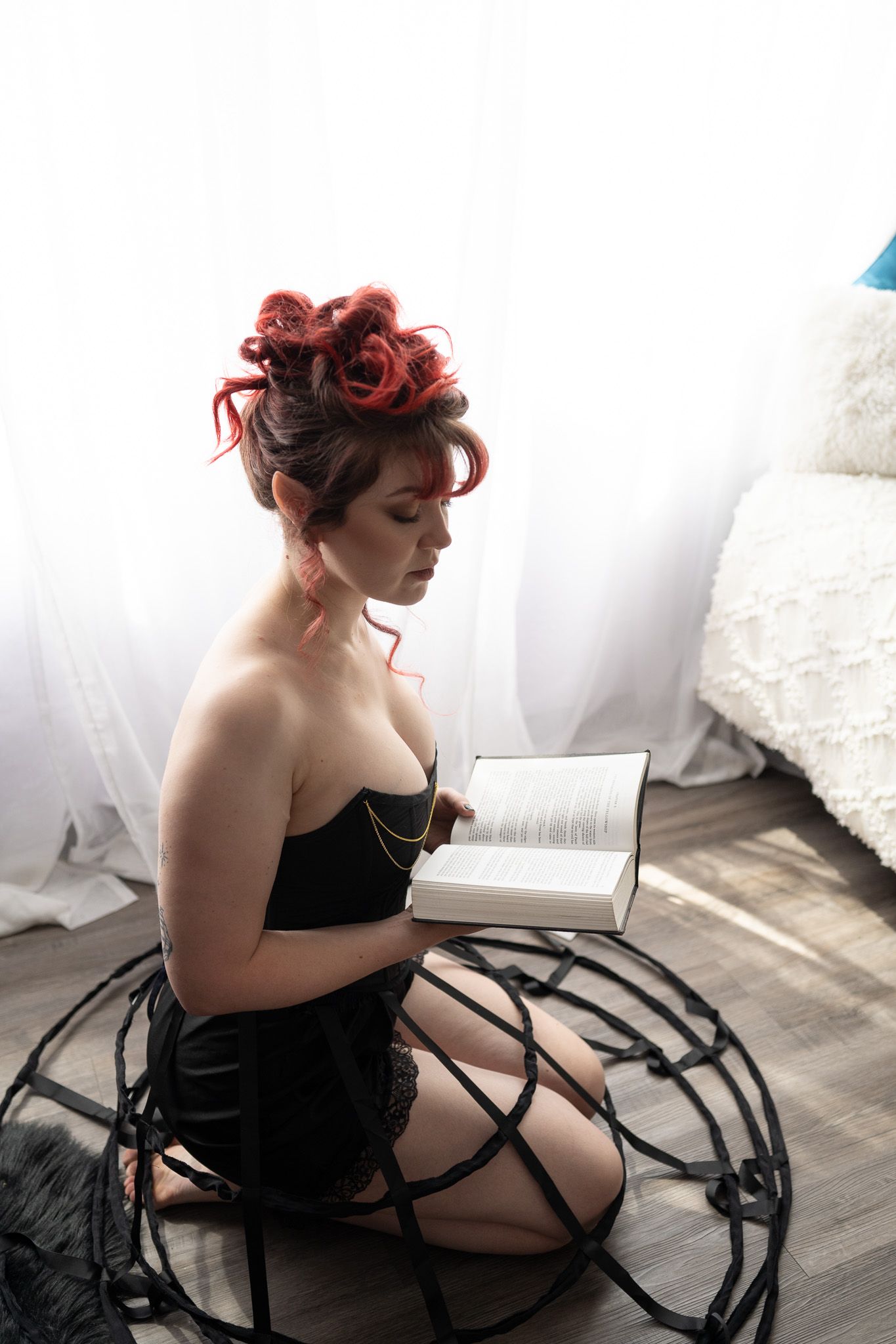 A woman in a boudoir photoshoot kneeling on the floor reading a book in a black corset and black hoop skirt. 