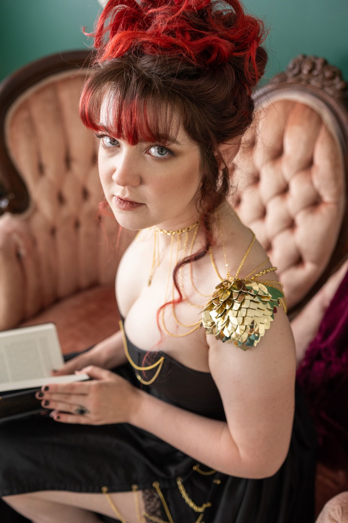 A woman looking over at the camera, holding a book. she is wearing a black dress and sitting on a pink couch. 