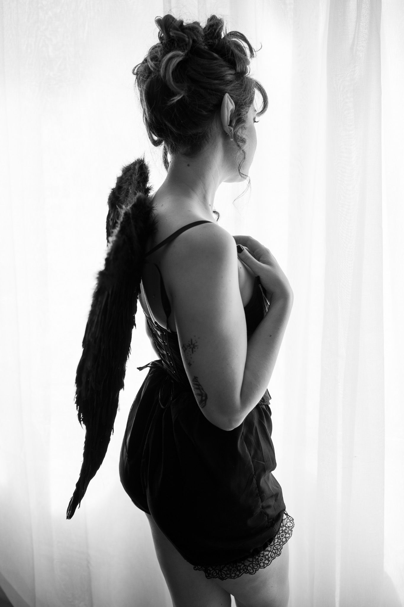 A woman in a black dress with angel wings is standing in front of a window.