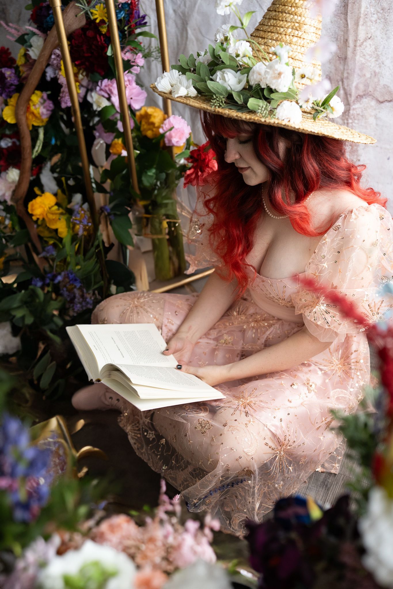 A woman in a witch hat is reading a book in a room surrounded by flowers.