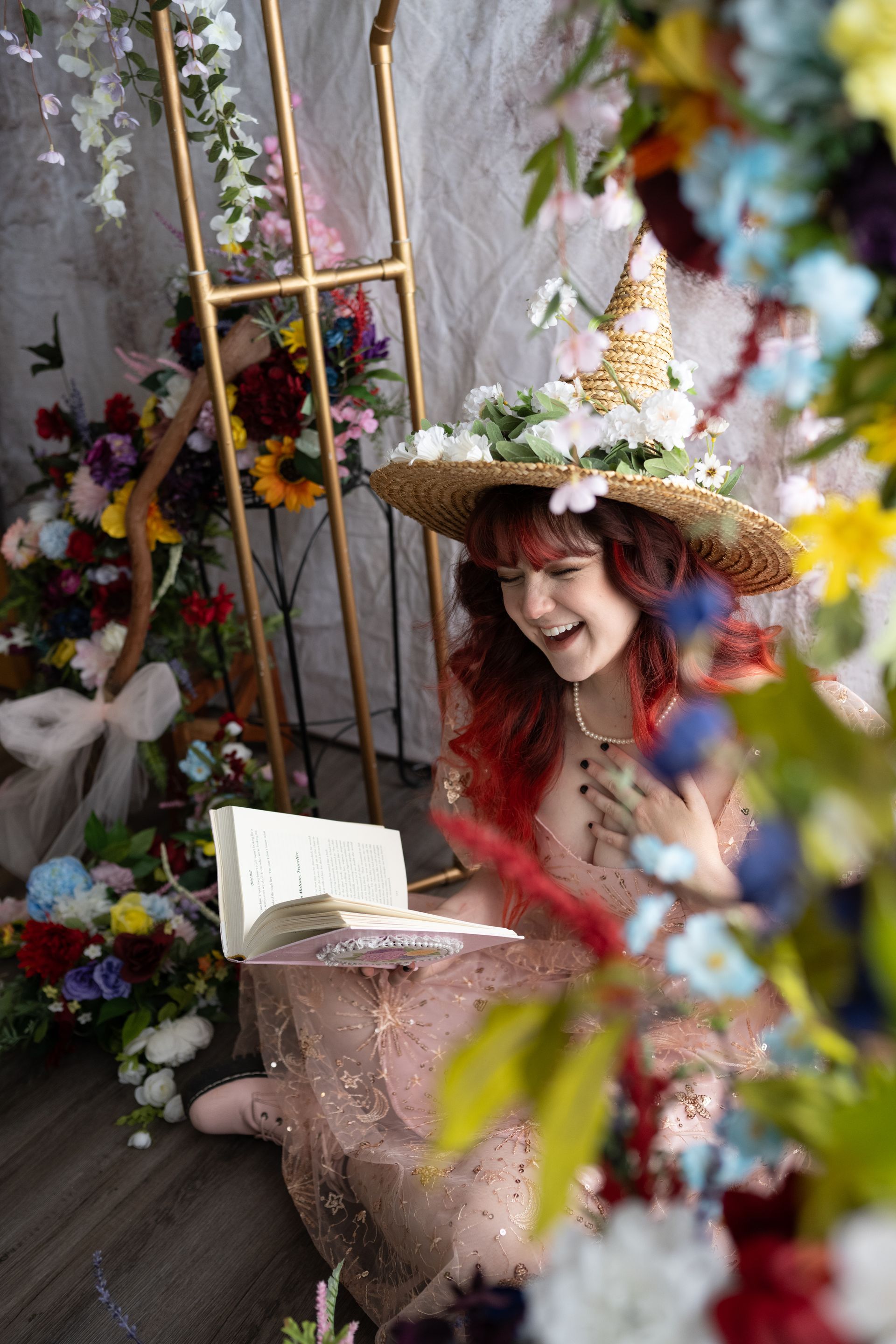 A woman in a witch hat is reading a book in a room surrounded by flowers.
