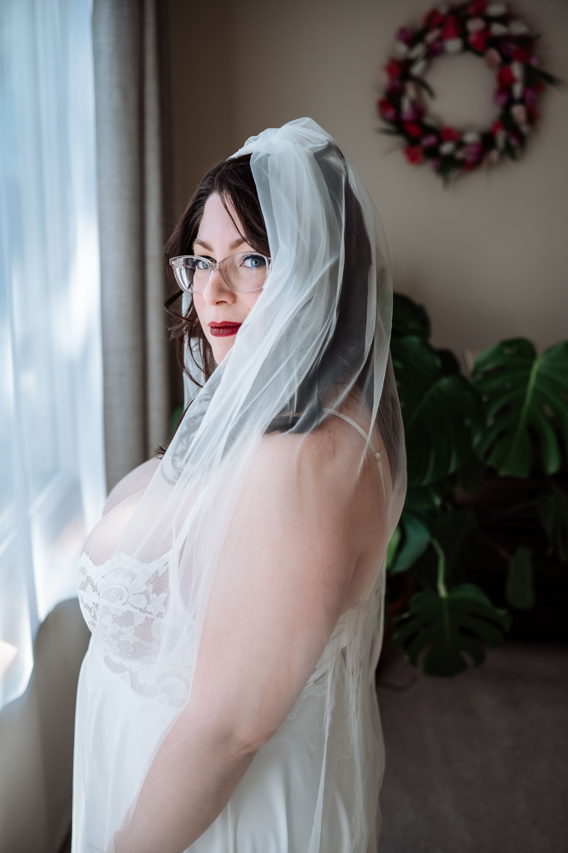 A woman in a wedding dress and veil is standing in front of a window.