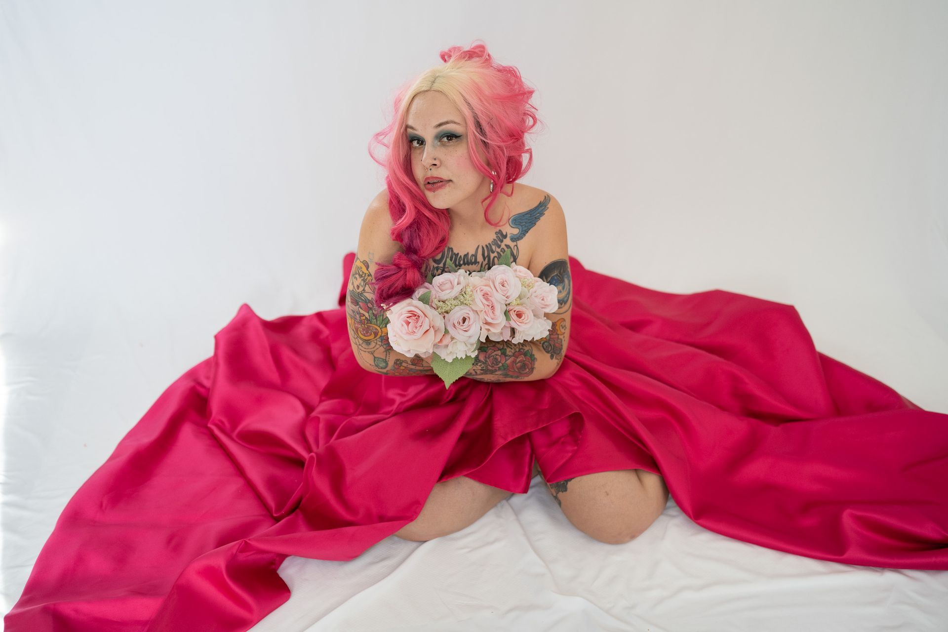 A woman with pink hair is sitting on a bed holding a bouquet of flowers.