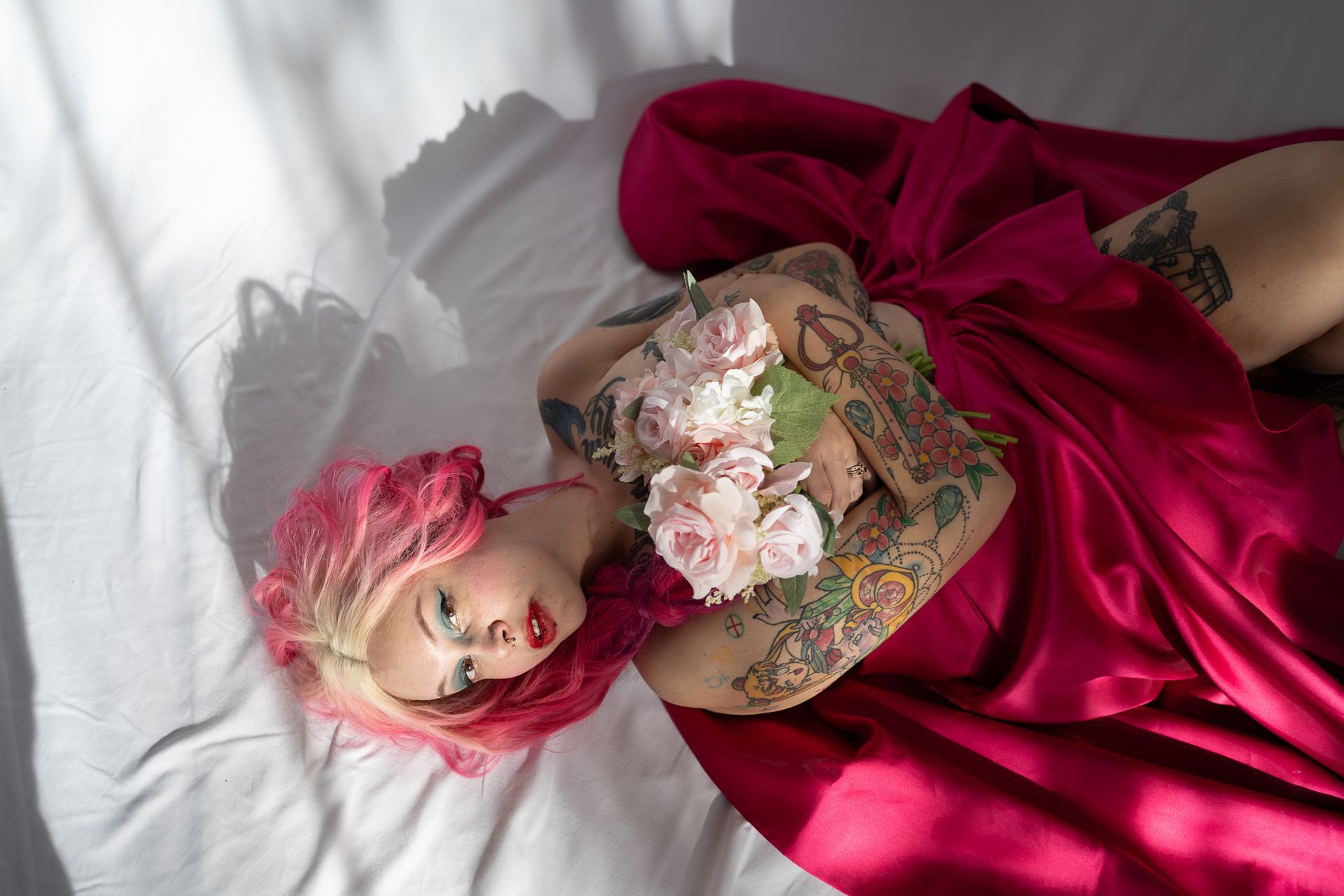 A woman with pink hair and tattoos is laying on a bed holding a bouquet of flowers.