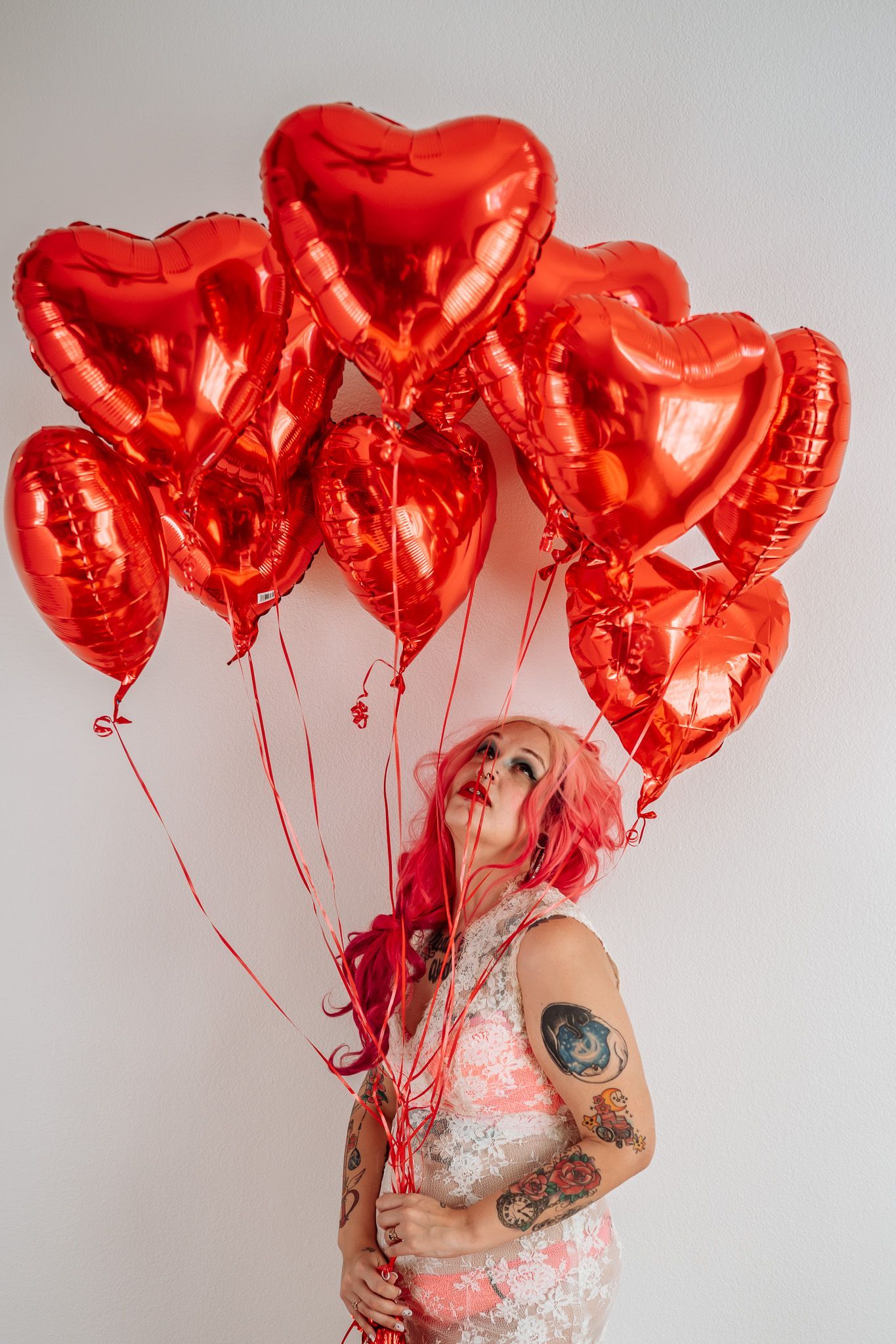 A woman with pink hair is holding a bunch of red heart shaped balloons.