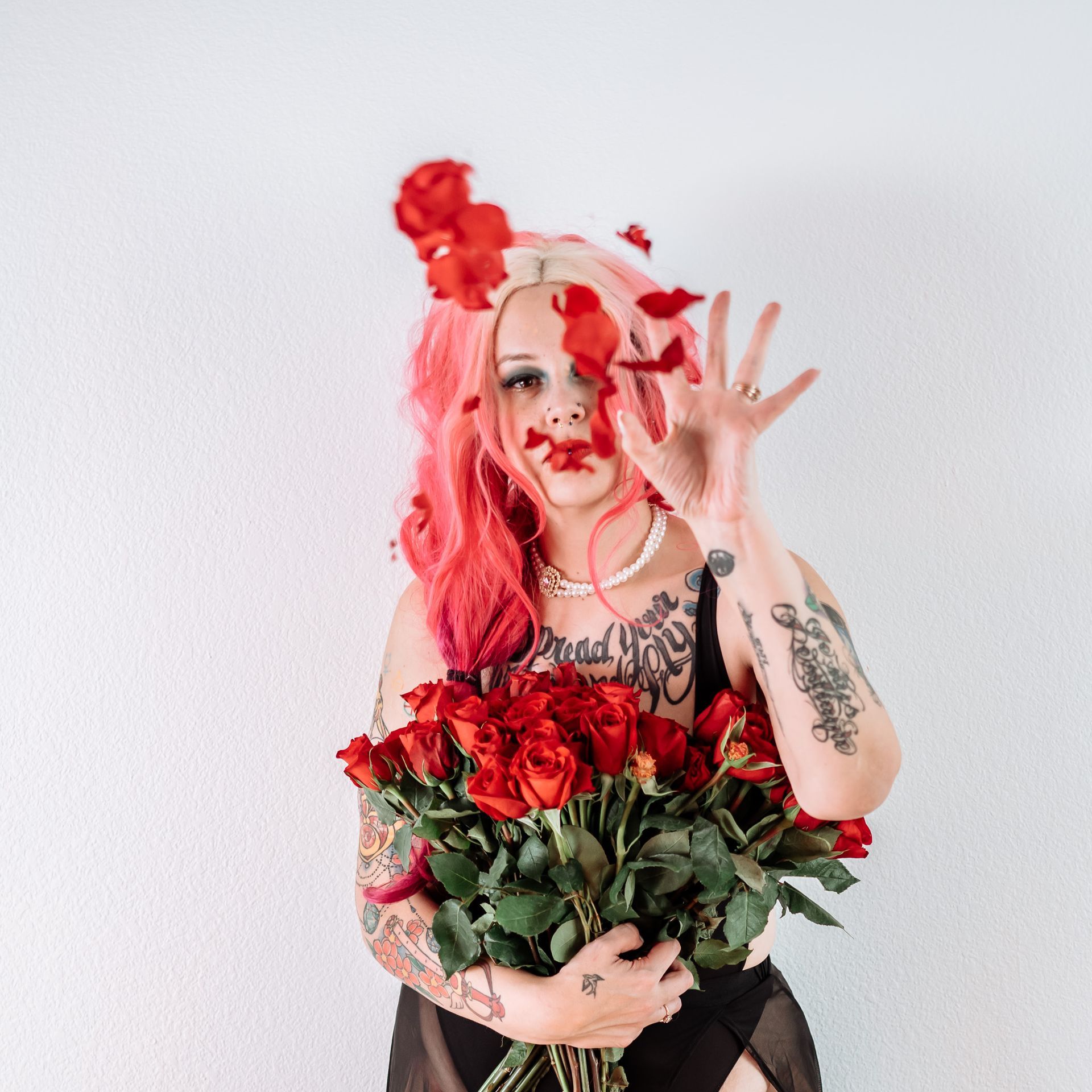 A woman with pink hair is holding a bouquet of red roses in front of her face.