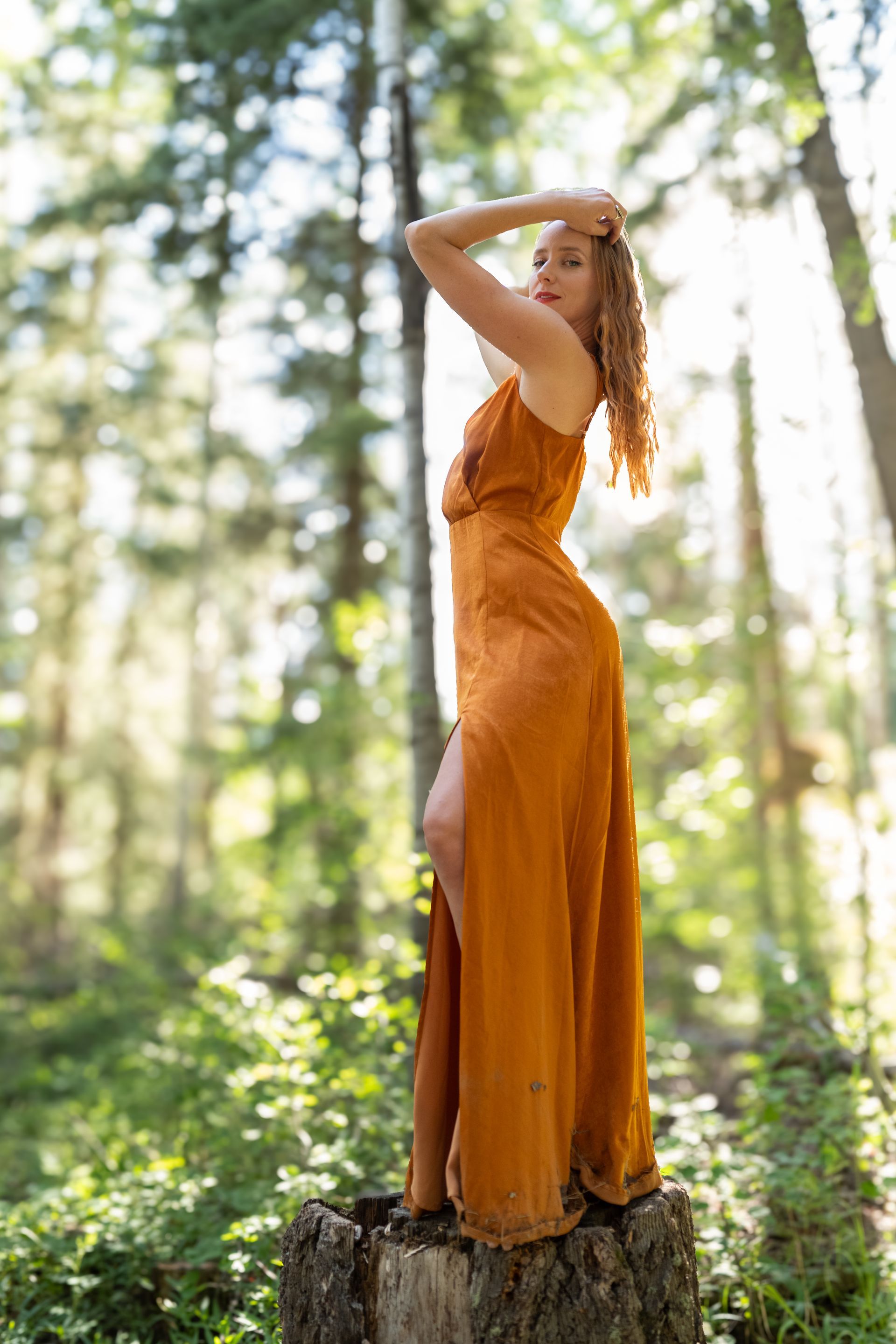 A woman in a long orange dress is standing on a tree stump in the woods.