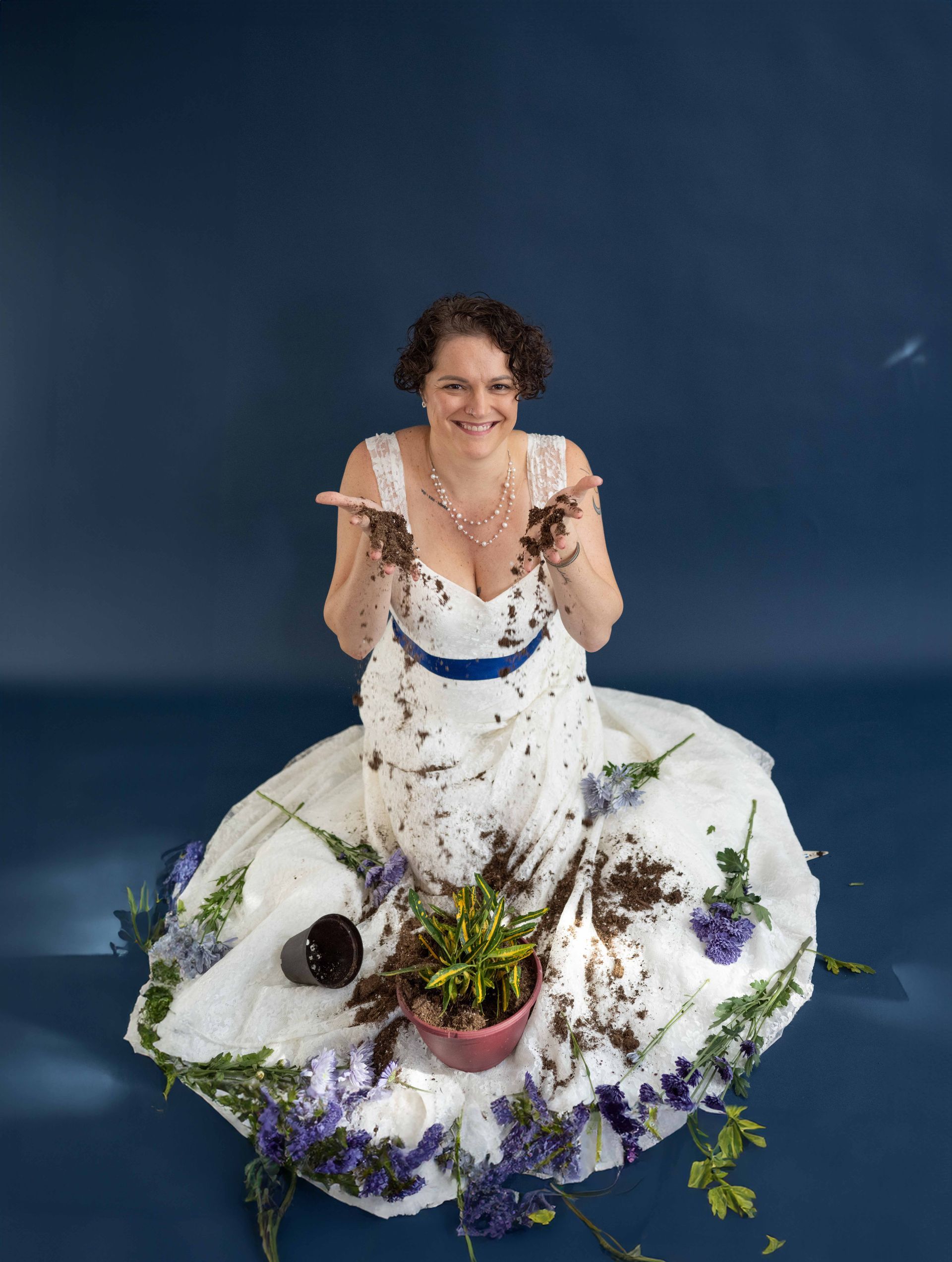 A woman in a wedding dress is sitting on the floor covered in flowers and dirt.