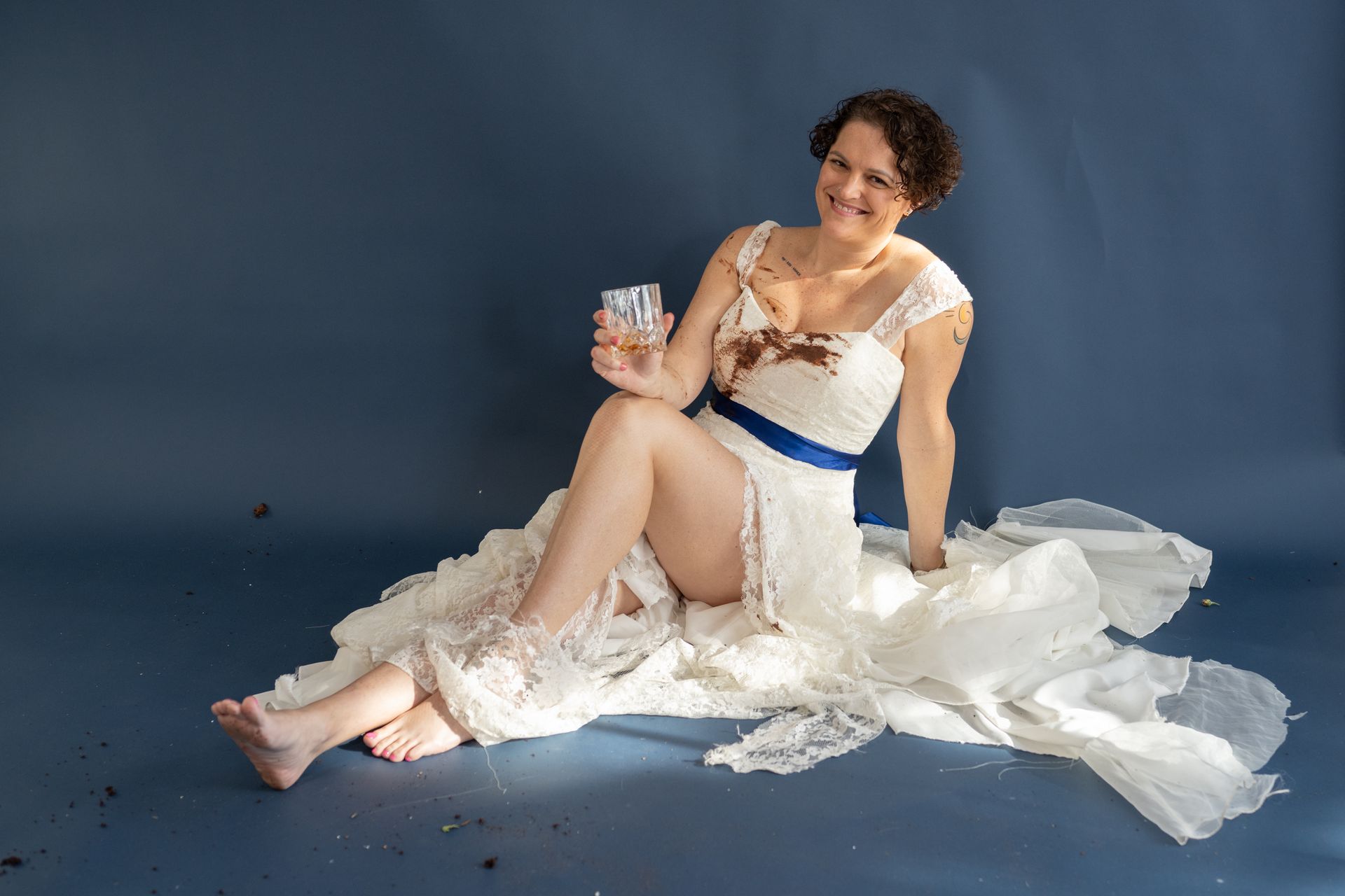 A photo of a woman sitting in a dirty wedding dress with a glass of whiskey on her knee.