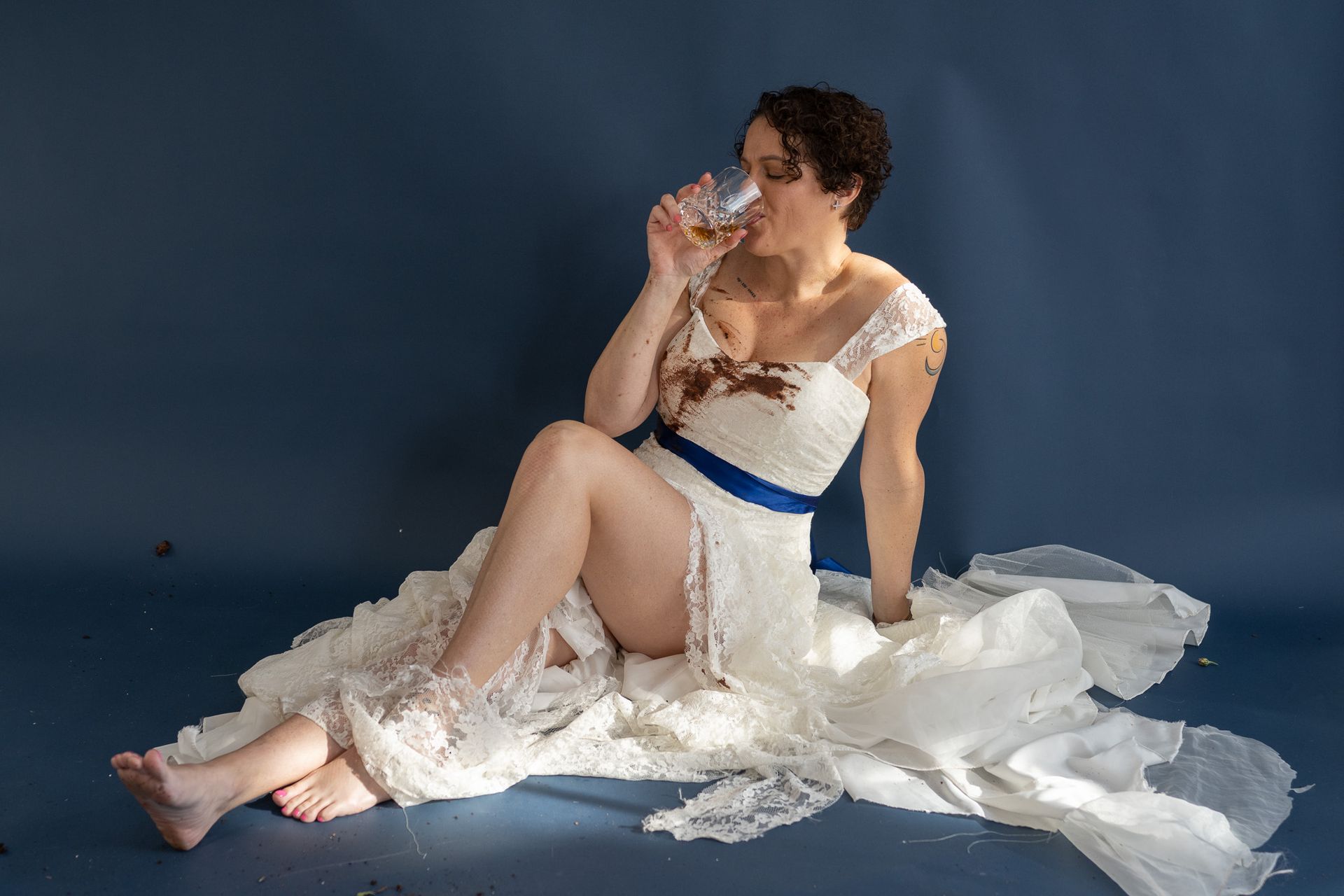 A woman in a wedding dress is sitting on the floor drinking from a glass.
