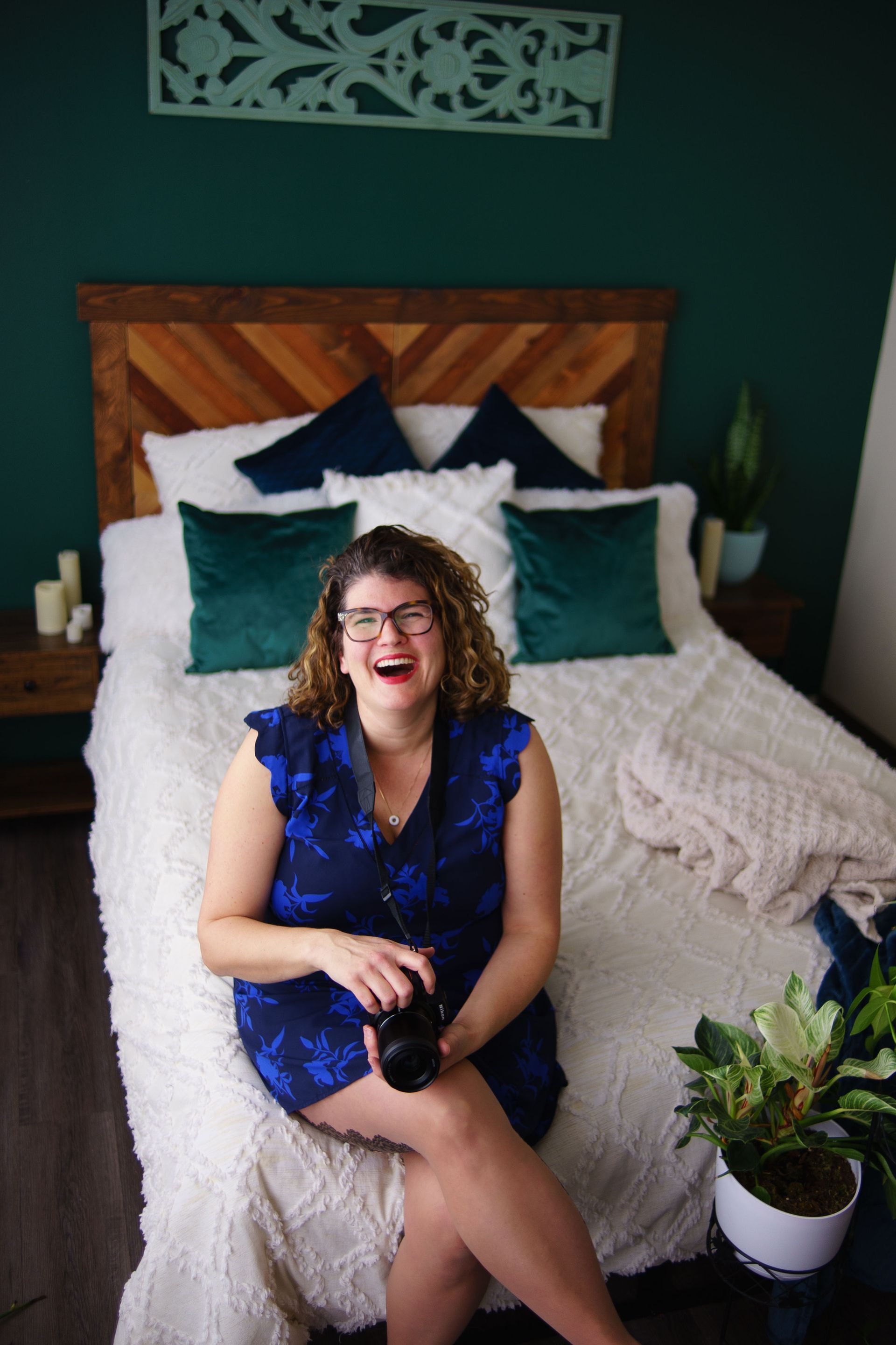A woman in a blue dress is sitting on a bed holding a camera.