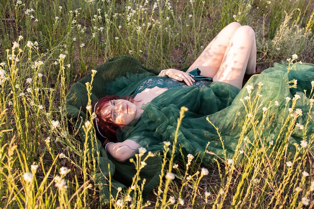 woman in an emerald green body suit laying down in tall grass, surrounded in white wild flowers. 