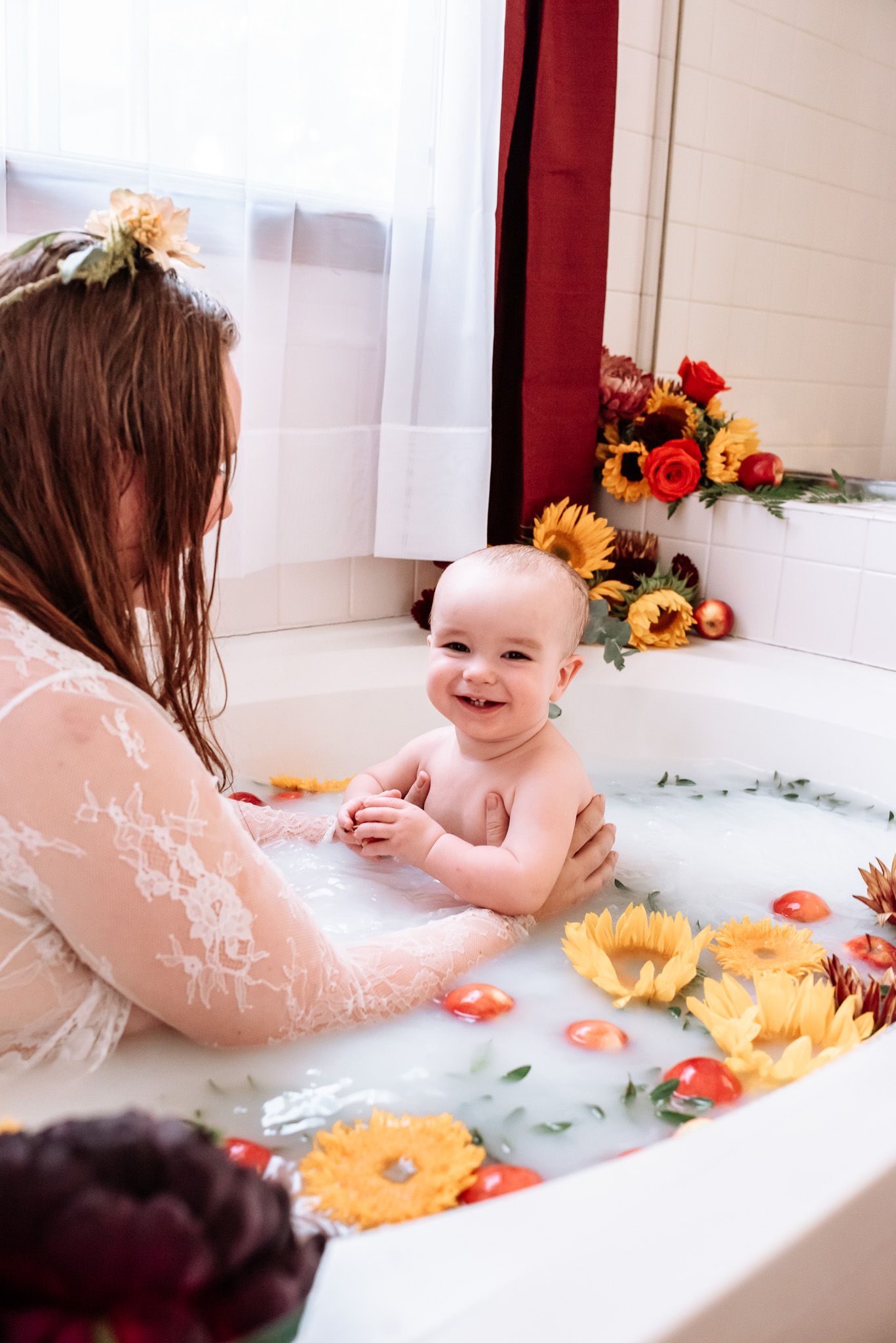 A woman is holding a baby in a bathtub filled with milk and flowers.