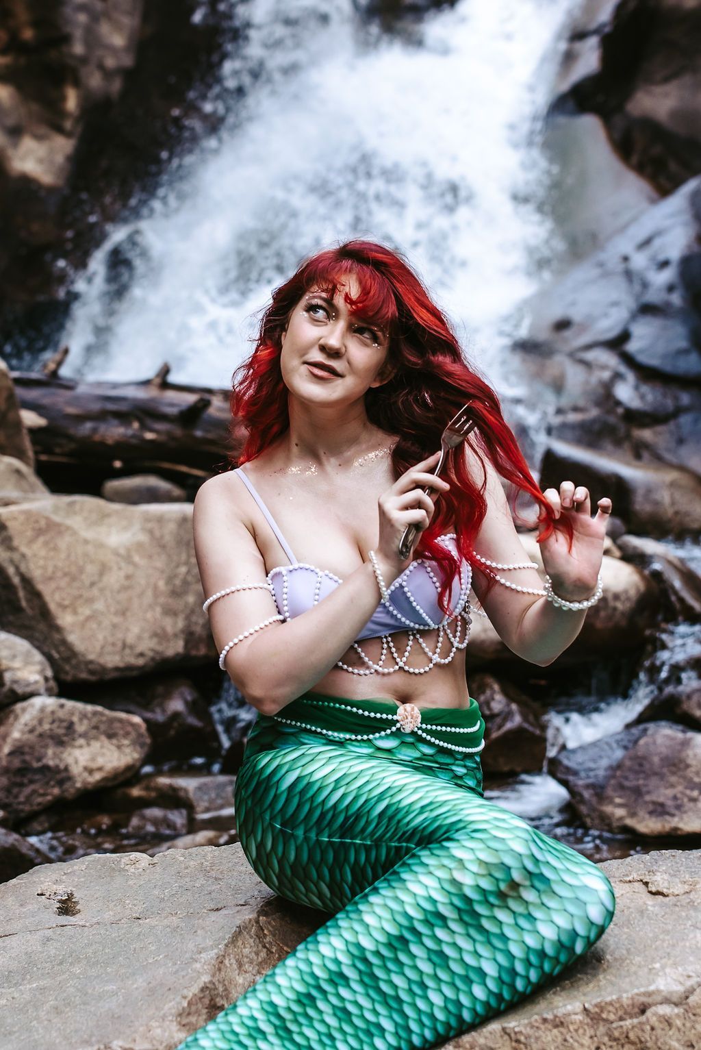 A woman dressed as a mermaid is sitting on a rock in front of a waterfall.