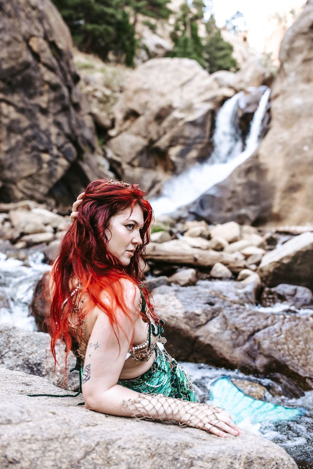 A woman dressed as a mermaid is sitting on a rock near a waterfall.