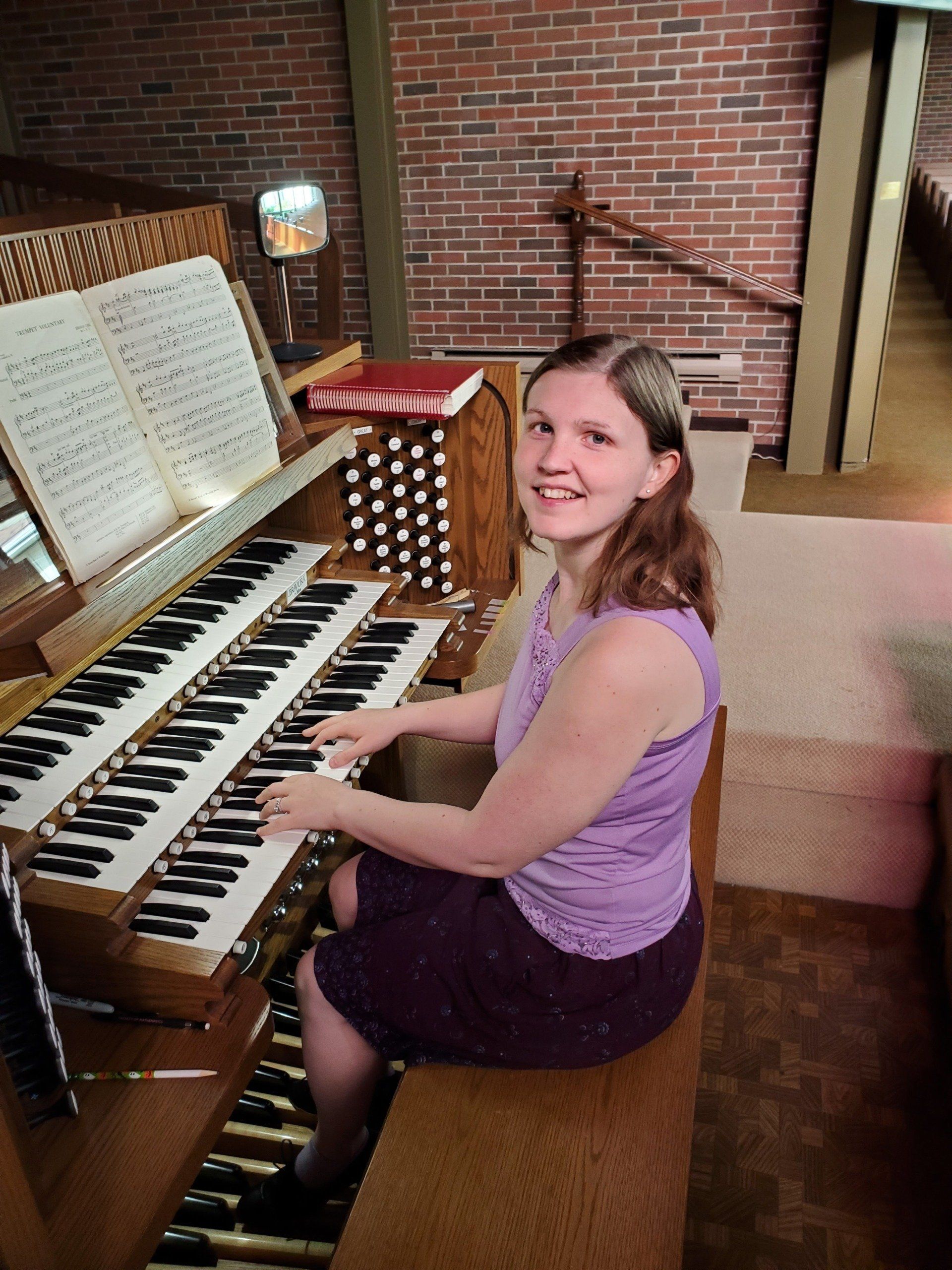 Woman playing an organ in a church, smiling. She wears purple and sits on a bench. Sheet music visible.
