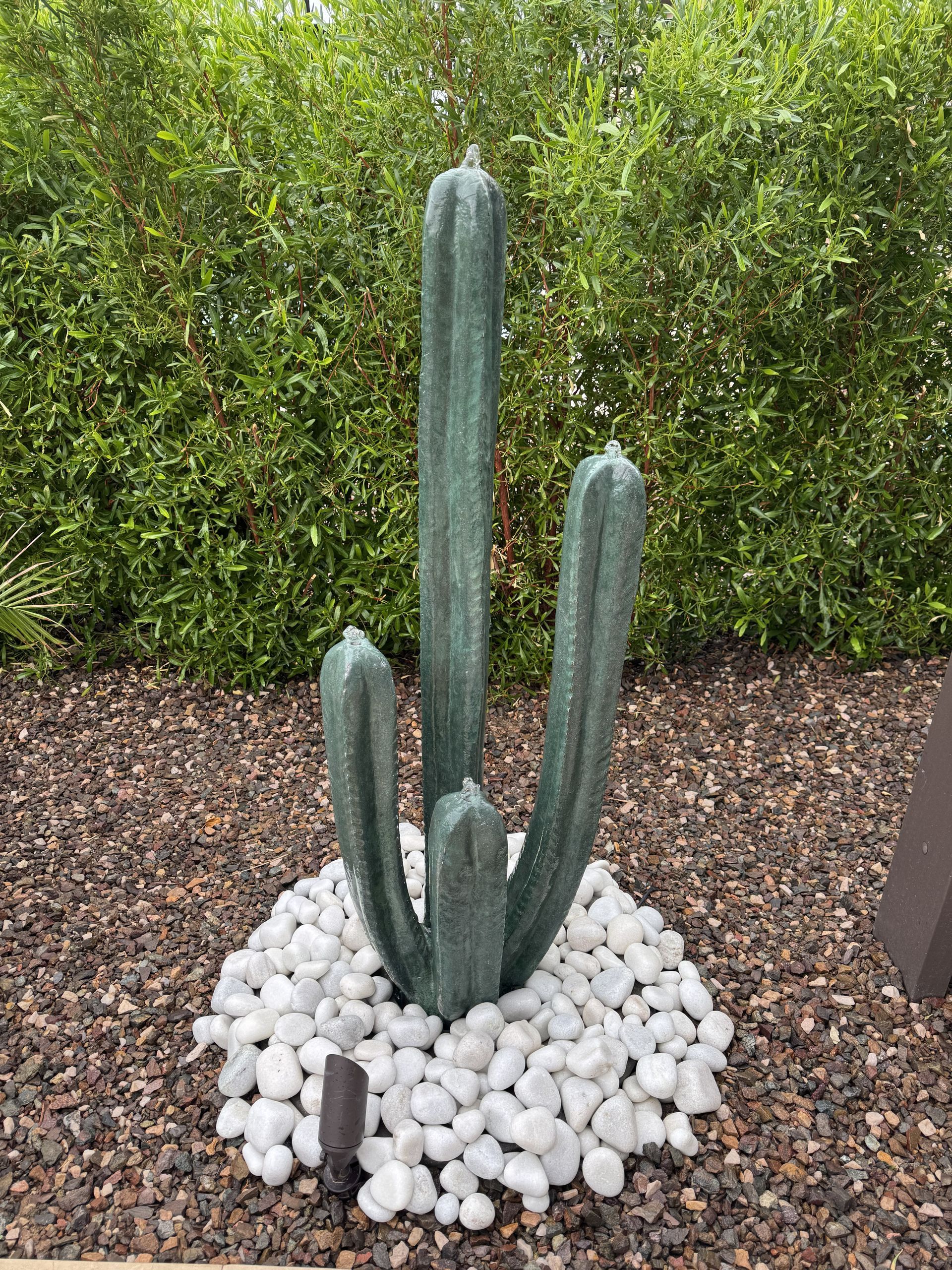 Green mosaic cactus sculpture surrounded by white pebbles, set against green shrubbery.