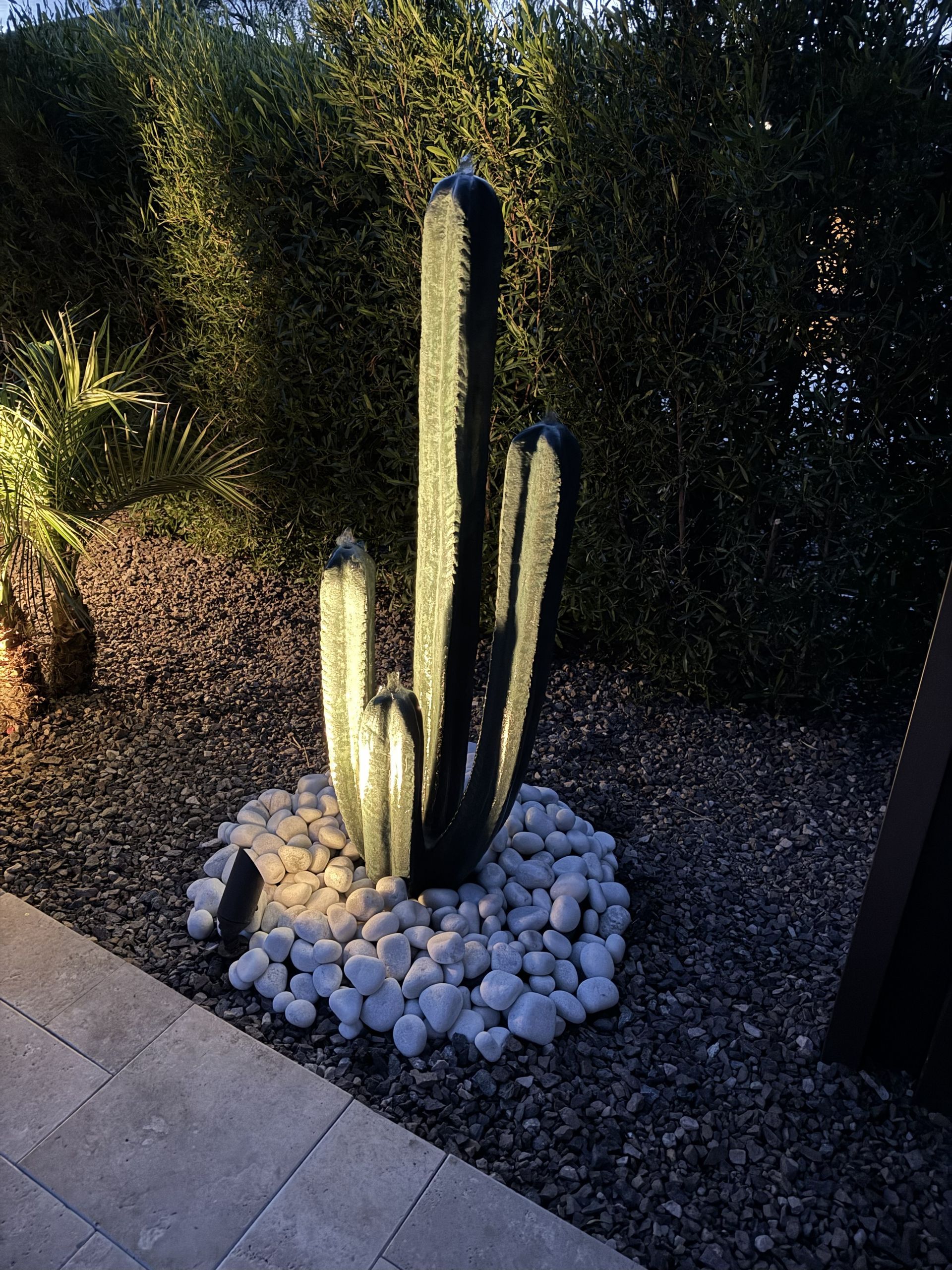 Lit-up cactus sculpture surrounded by white stones and gravel in a garden at night.