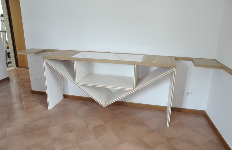 Tavolo espositivo pieghevole bianco con ripiani e supporto triangolare, posizionato contro una parete liscia su un pavimento piastrellato.