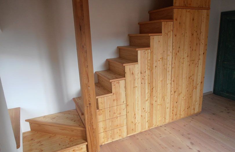 Scala in legno con pannellatura verticale in una stanza semplice con pavimento in legno chiaro e pareti bianche.