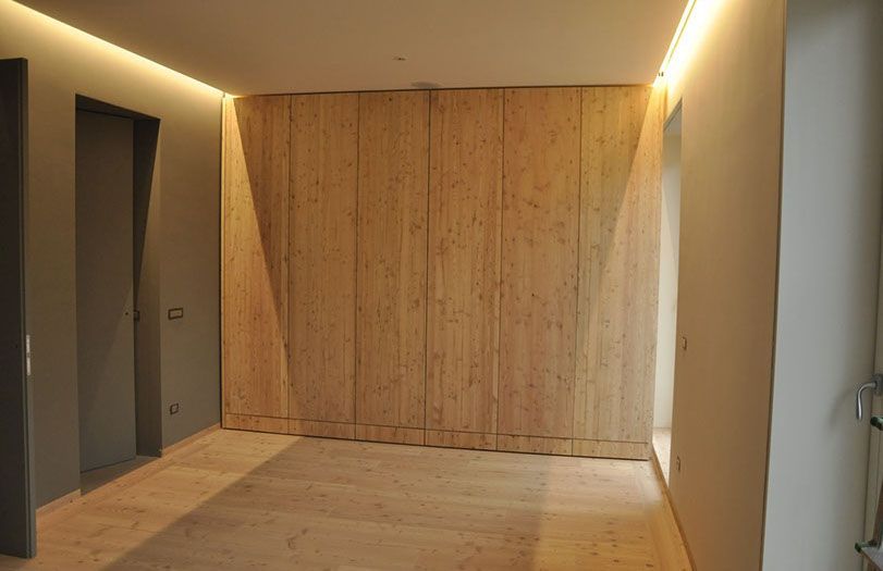 Stanza moderna vuota con pareti e pavimento in legno chiaro, illuminazione a incasso e porte grigie sulla sinistra.