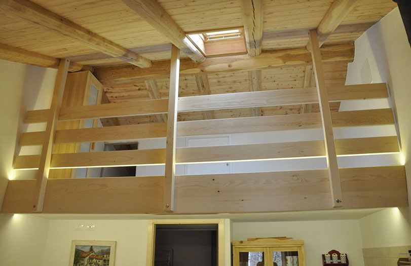 Soppalco in legno con ringhiere orizzontali sopra un corridoio, illuminato da calde applique.