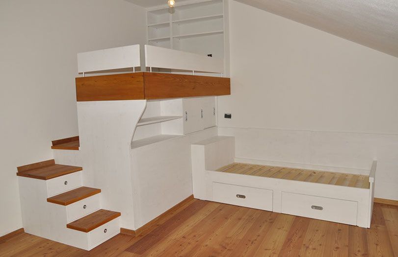 Piccola stanza soppalcata con scala in legno, armadi a muro bianchi e un divano letto basso sotto il soffitto inclinato.