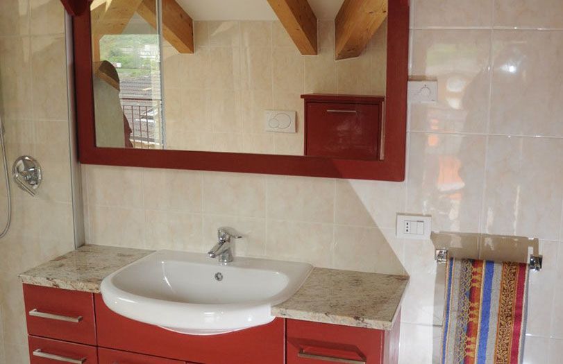 Mobile da bagno con lavabo bianco, ante rosse e un grande specchio su una parete rivestita di piastrelle beige.