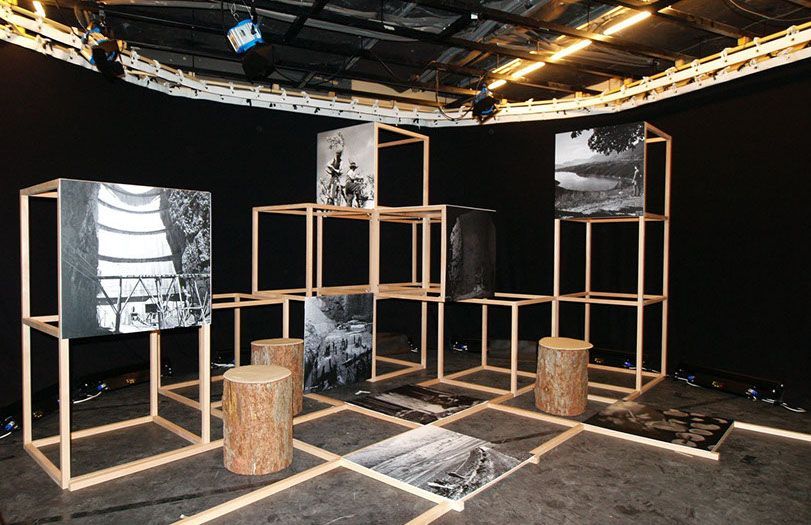 Installazione artistica con cornici cubiche in legno, pannelli in bianco e nero, tronchi d'albero e tappeti in una stanza buia.