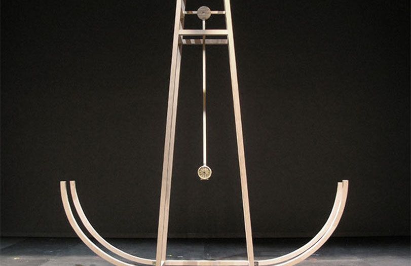 Scultura astratta in legno con elementi laterali curvi e pesi metallici sospesi su sfondo nero.