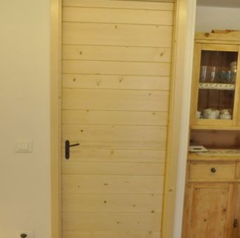 Porta interna in legno chiaro con maniglia nera accanto a un mobile in legno in una stanza dai toni chiari.