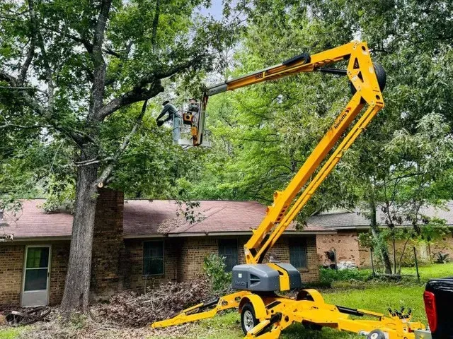 A worker in a yellow lift trimming a tree near a brick house on a sunny day.