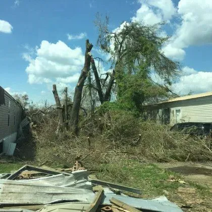 A damaged tree and debris next to a mobile home after a storm, under a partly cloudy sky.