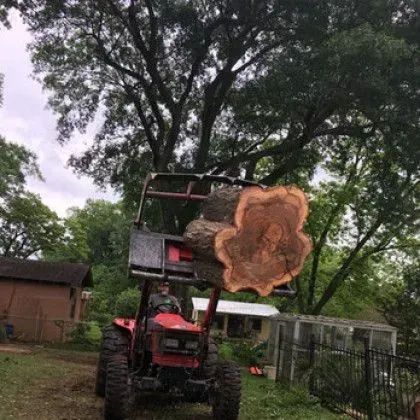 Man operating red tractor lifts a large tree trunk outdoors, near a house.
