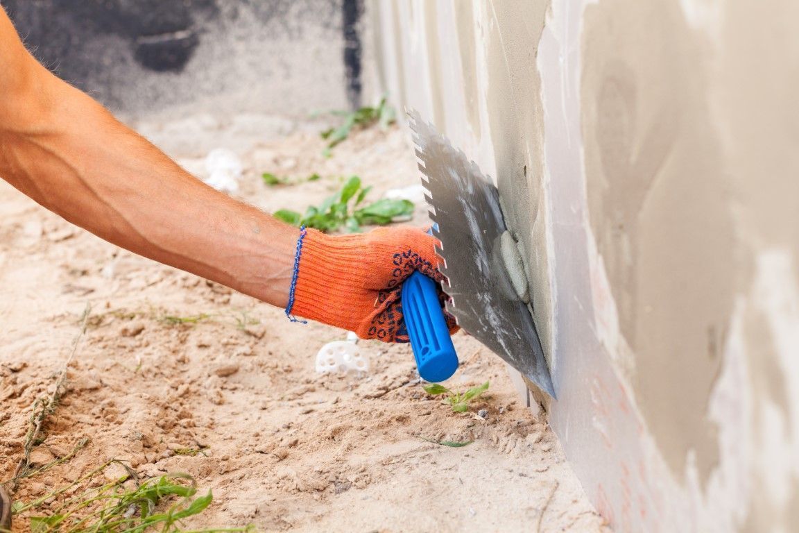 Person with orange glove applying waterproofing sealant to a wall with a blue tool