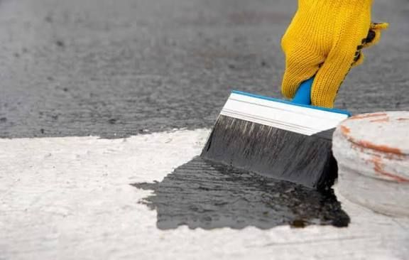 A gloved hand paints a white surface with black paint using a brush.