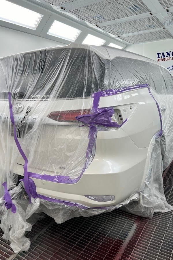 White car rear in a paint booth, covered in plastic masking with purple tape around the tail light and bumper area