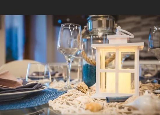 Apparecchiatura della tavola in tema costiero: lanterna con candele, bicchieri da vino, decorazioni blu e beige.