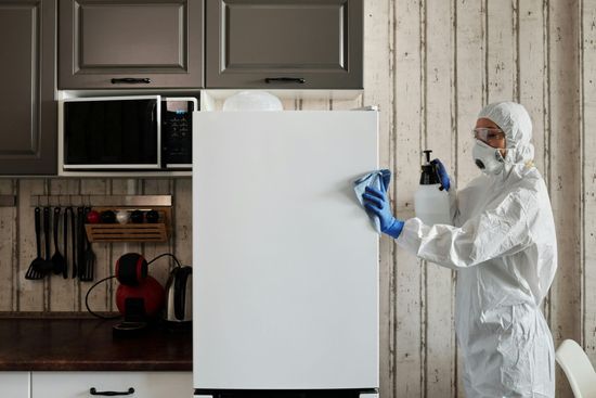 Persona in tuta protettiva che disinfetta un frigorifero bianco in cucina.