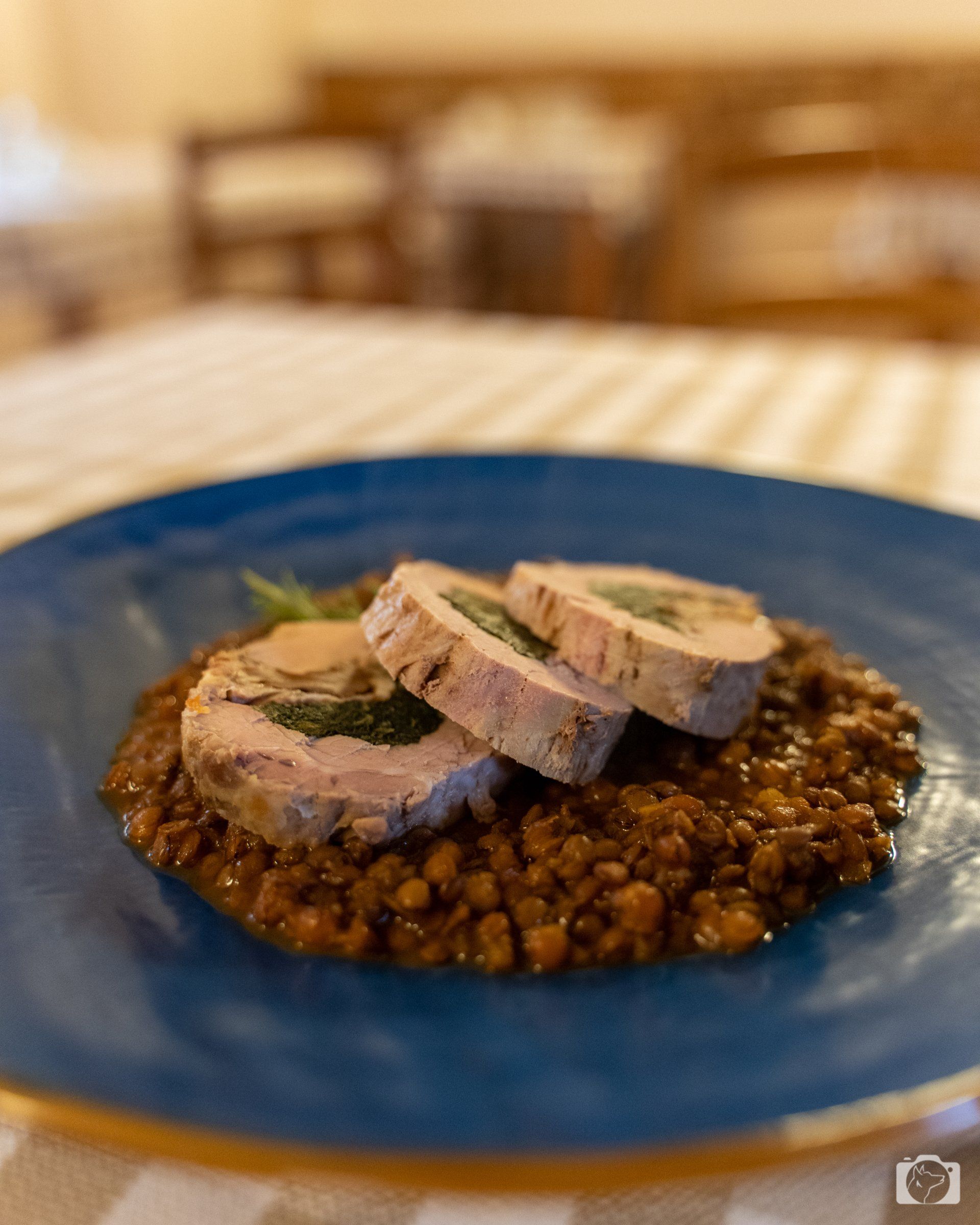 Rotolo di carne affettata su un letto di lenticchie, presentato su un piatto blu con sfondo sfocato.