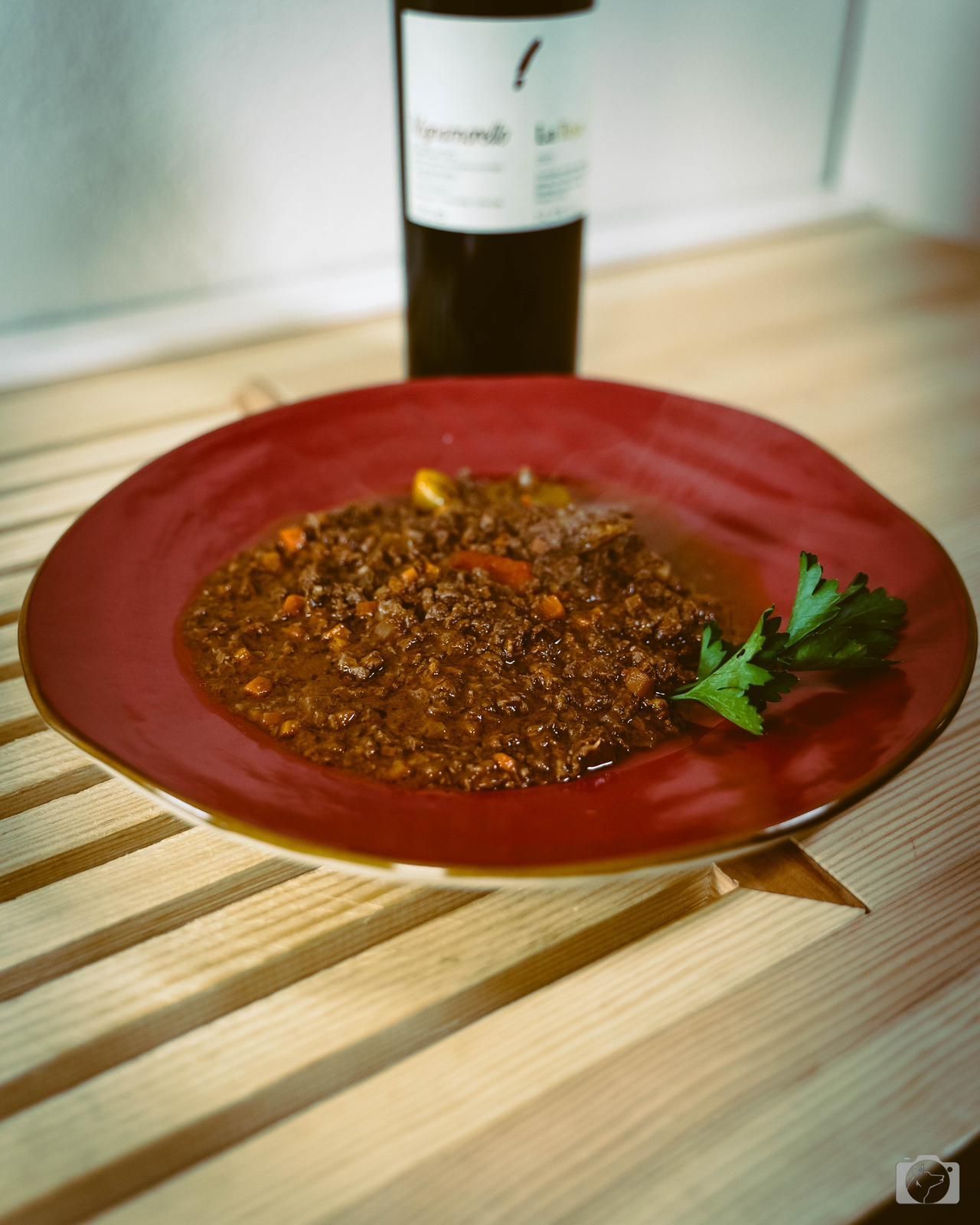 Piatto di salsa scura con guarnizione di prezzemolo; bottiglia di vino sullo sfondo su una superficie di legno.